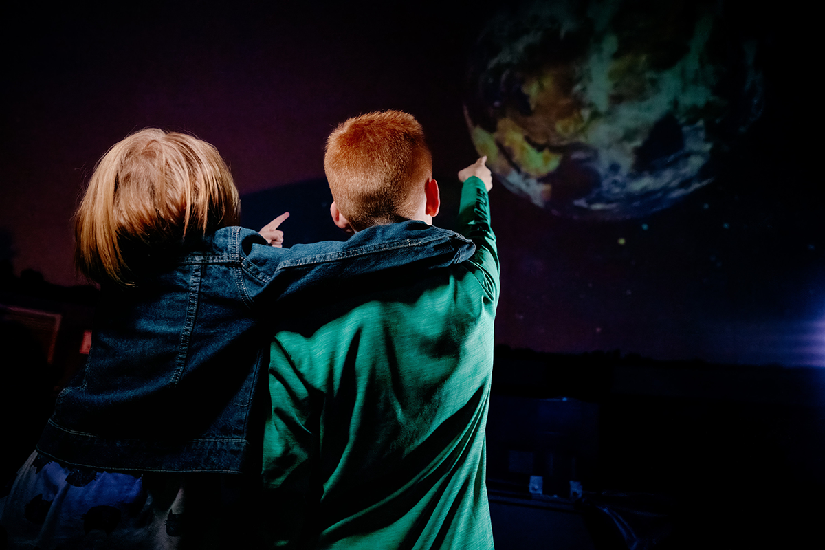 2 children with backs turned to camera pointing at planet in planetarium